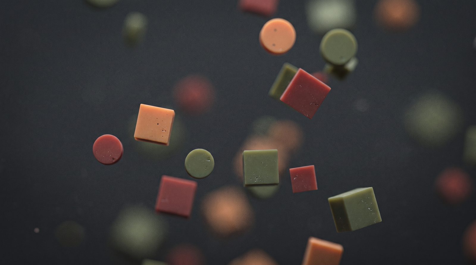 Matte green, orange and red discs and cubes floating apart in mid-air against a dark background